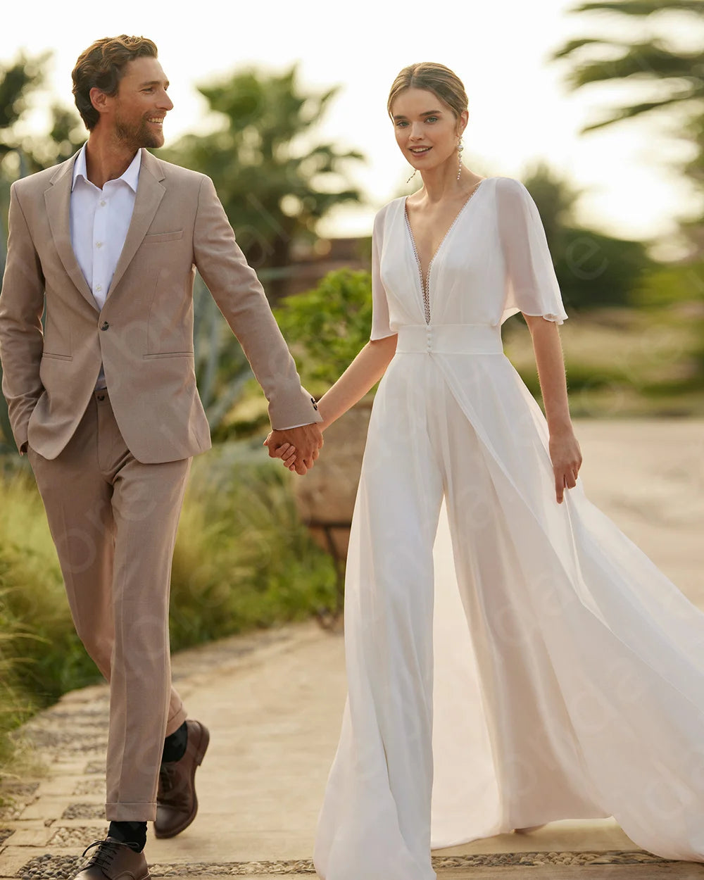 Bride wearing a white two-piece boho wedding jumpsuit with an elegant sheer chiffon half-sleeve top layer, walking and holding hands with a groom in a tan linen suit.