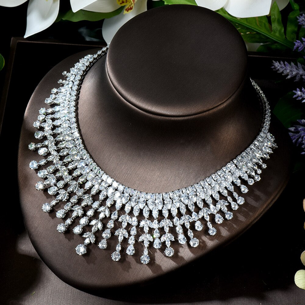 Direct view of the multi-row clear crystal bridal bib collar necklace showcased on a contoured dark brown display bust, highlighted by white orchid blossoms in the background.