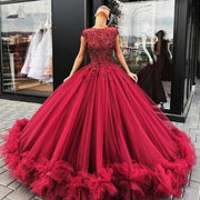 A model posing elegantly outdoors in a long, dark red ball gown featuring intricate crystal beading and a voluminous tulle skirt with ruffled hem details.
