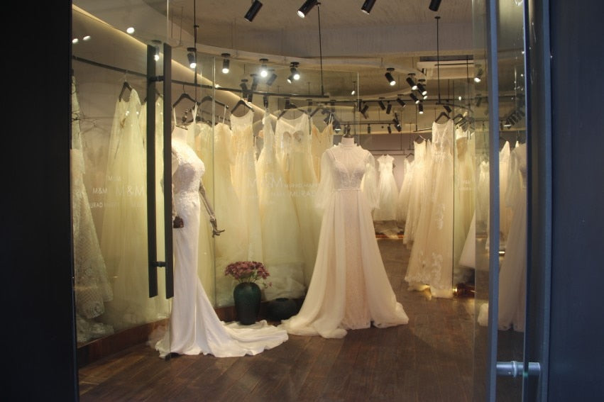 A view through a glass storefront into a bridal boutique featuring multiple mannequins displaying various elegant wedding gowns on hangers.