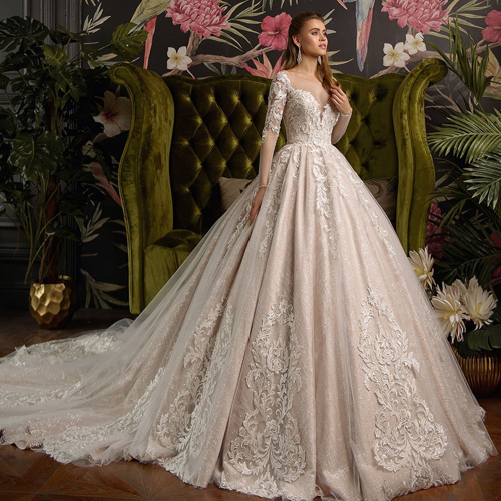 A gorgeous, shiny ball gown wedding dress featuring long lace sleeves, a deep V-neckline, and an expansive chapel train, set in an elegant room with a green velvet sofa.