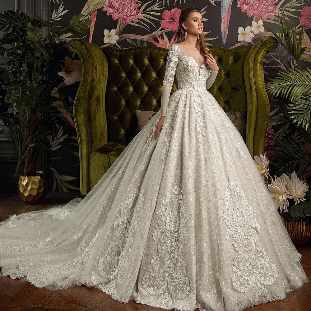 Full view of the bride posing in a gorgeous, shiny ball gown wedding dress with long lace sleeves and a chapel train against a botanical wallpaper background.