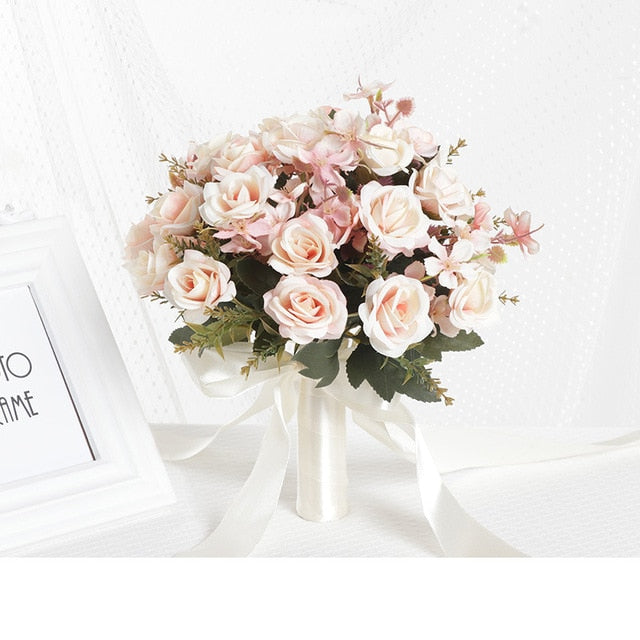 Front look at a romantic dusty pink artificial bridal bouquet composed of soft pink and ivory silk roses arranged with small matching ribbons on a texturized white fabric backdrop.