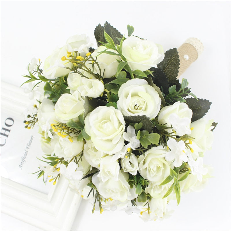 High-angle product photograph of a white handmade wedding bridal bouquet composed of silk ivory roses, green foliage, and tiny yellow flower sprigs, styled with a burlap and lace wrapped handle.