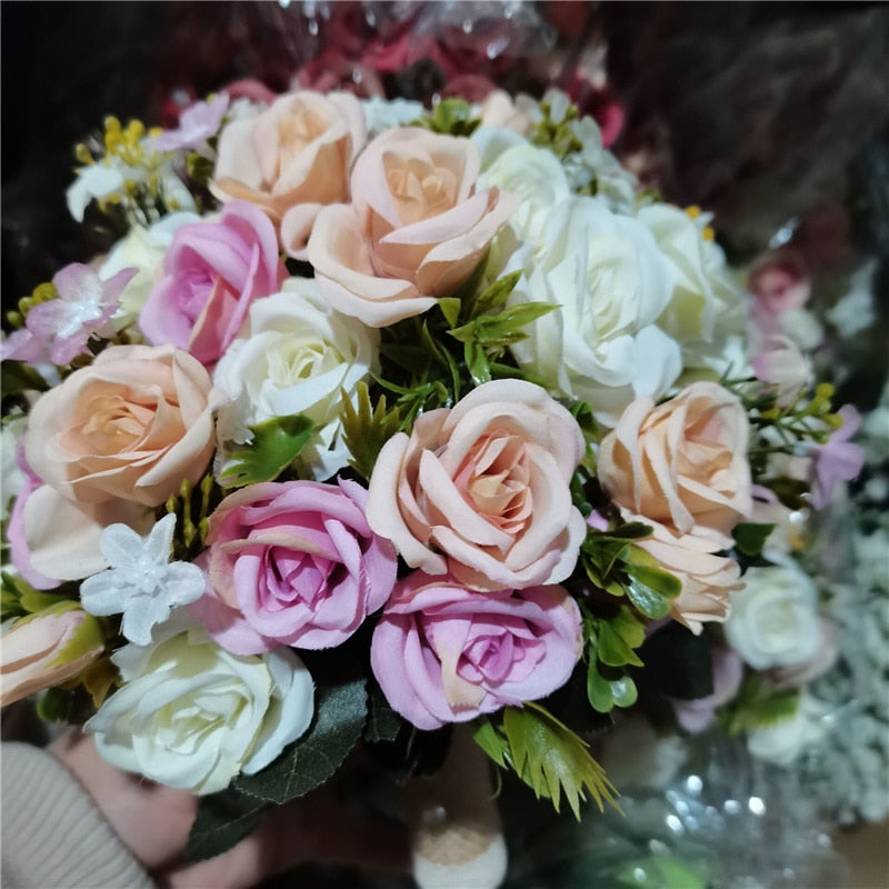 Close-up top view of a mixed faux rose wedding bouquet featuring tightly clustered silk roses in shades of pastel pink, light peach, and creamy white, accented with deep green leaves.