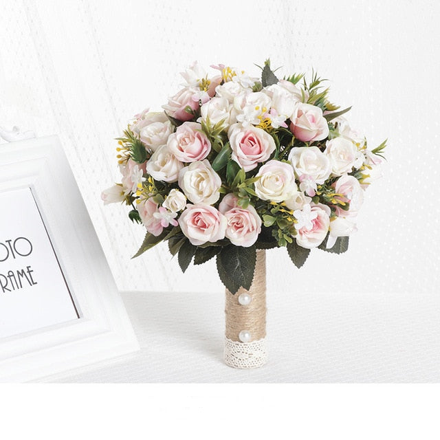 Studio product display of a round artificial bridesmaid bouquet packed with soft pink and ivory silk roses, built with a decorative burlap and lace handle next to a white photo frame.