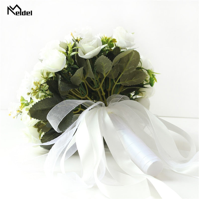 Low-angle view looking up at the base of an all-white artificial bridal bouquet, showcasing the neatly tied green plastic stems bound together with wide, trailing white satin and sheer organza ribbons.