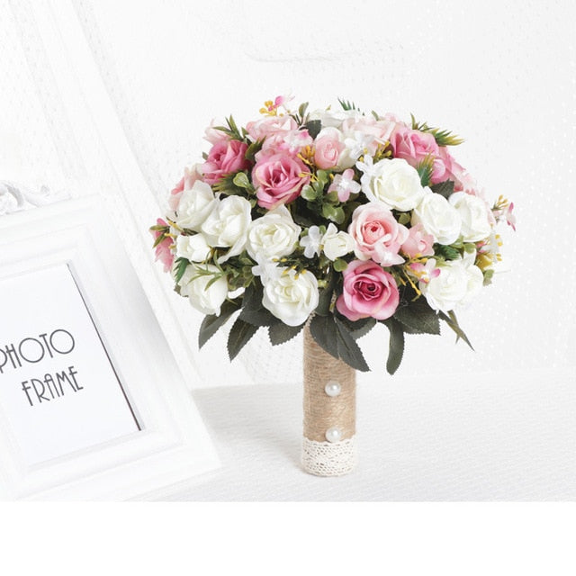 Brightly lit product display of a round artificial wedding bouquet showcasing coral, peach, and deep pink silk roses, built with a decorative burlap and lace stem wrap next to a white photo frame.