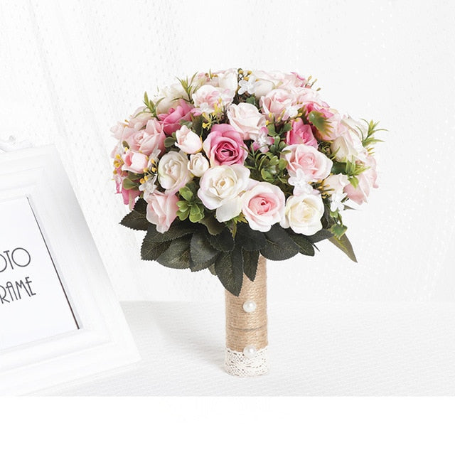Symmetrical front look at a romantic dusty pink artificial bridal bouquet composed of soft pink and ivory silk roses arranged with small matching ribbons on a texturized white fabric backdrop.