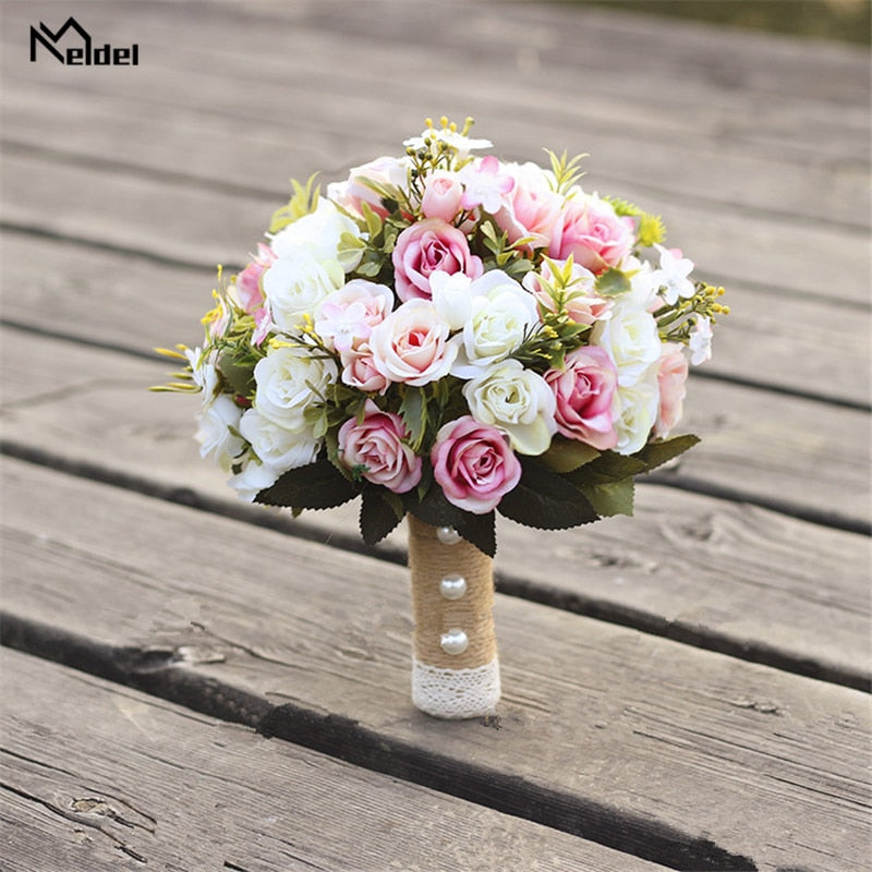 Studio product shot of a Meldel artificial flower wedding bouquet featuring pink and white silk roses, styled with a burlap-wrapped handle accented by pearl pins and lace trim on a rustic wooden surface.