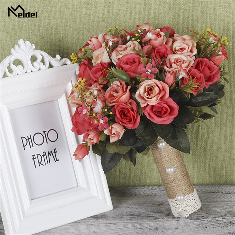 Product view of a vibrant coral and bright pink artificial bridesmaid bouquet packed with realistic silk roses, styled with a natural twine handle decorated with white pearls next to a white picture frame.