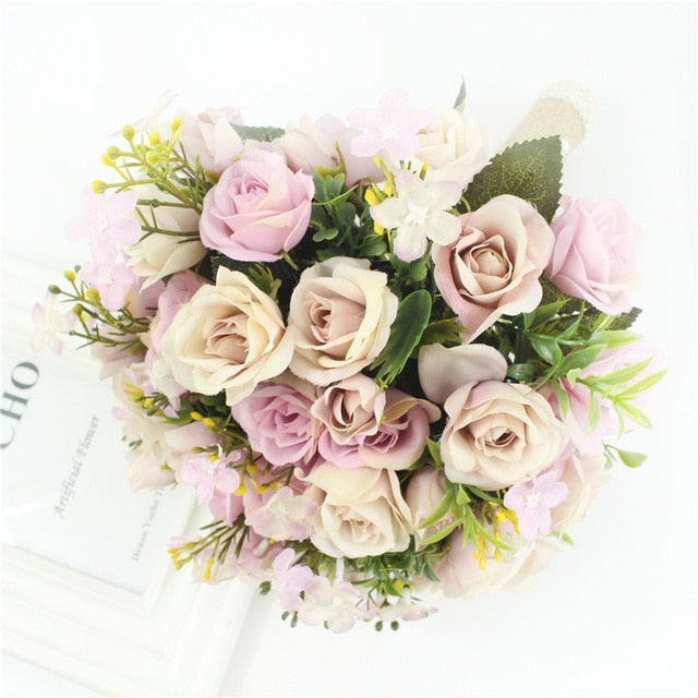 Top-down flat lay view of a faux bridal flower arrangement displaying muted mauve, light purple, and cream-colored silk roses mixed with small green foliage accents.