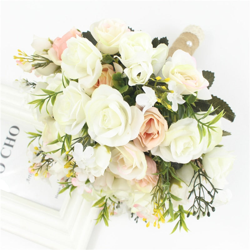 High-angle product photograph of a white handmade wedding bridal bouquet composed of silk ivory roses, green foliage, and tiny yellow flower sprigs, styled with a burlap and lace wrapped handle.