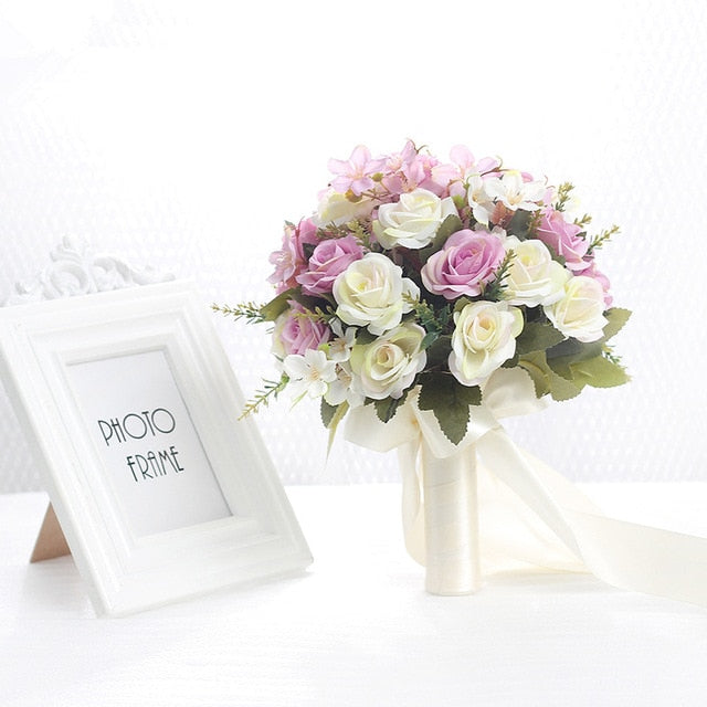 Elegant studio product shot of a minimalist cream-colored faux wedding bouquet featuring silk white roses, delicate green sprigs, and long flowing ivory satin ribbons.
