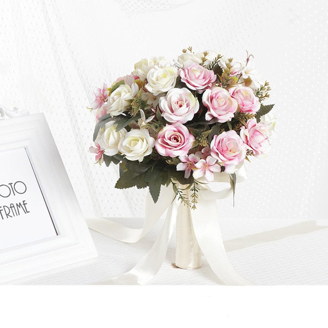 Front view of a round faux rose bridal wedding bouquet featuring cream and soft pink silk blossoms, styled with an ivory satin ribbon handle next to a white picture frame.