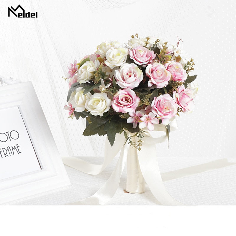 Front view of a round artificial bridal bouquet displaying mixed white, champagne, and blush pink silk roses, constructed with an ivory satin ribbon handle next to a white picture frame.