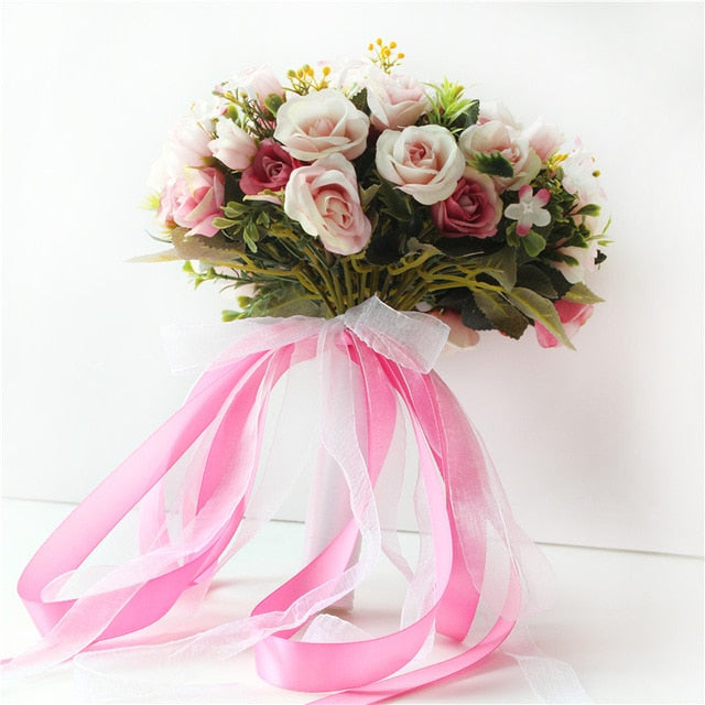 Studio product shot of a vibrant artificial flower bridal bouquet featuring a cascade of trailing white and bright pink satin ribbons attached to the base of mixed silk roses.