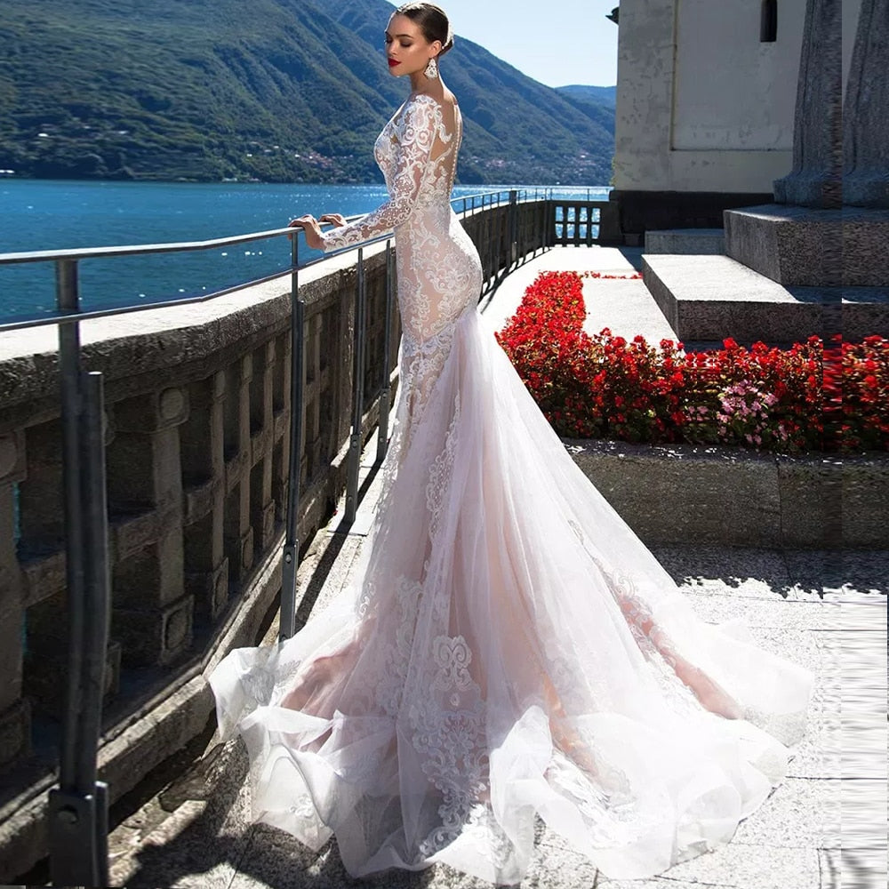 Elegant side profile view of a bride in a long-sleeve mermaid bridal gown with a dramatic lace train, posing on a balcony overlooking the water.