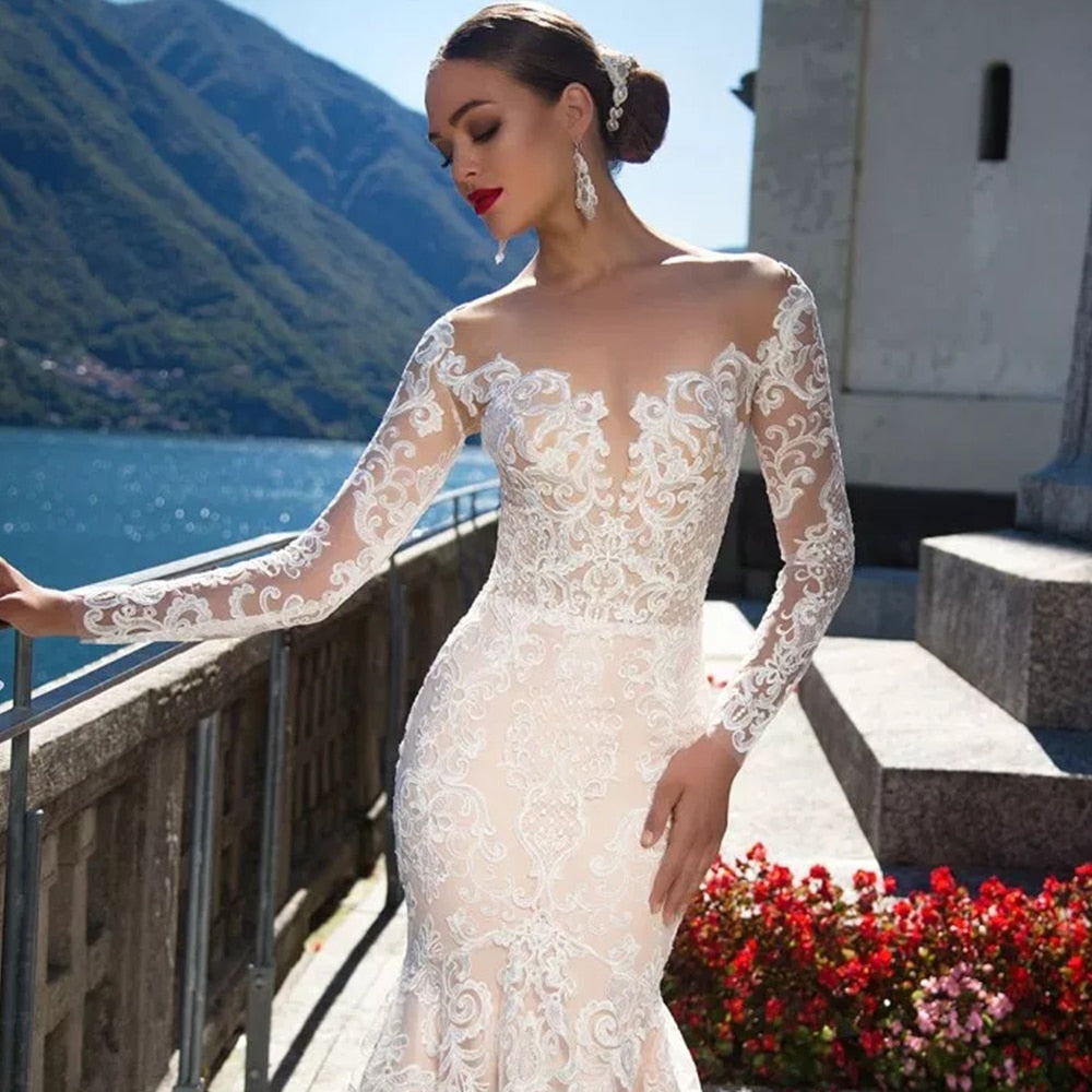 Close-up front view of a bride wearing a sexy long-sleeve mermaid wedding dress with delicate lace floral appliques and a sheer illusion neckline.