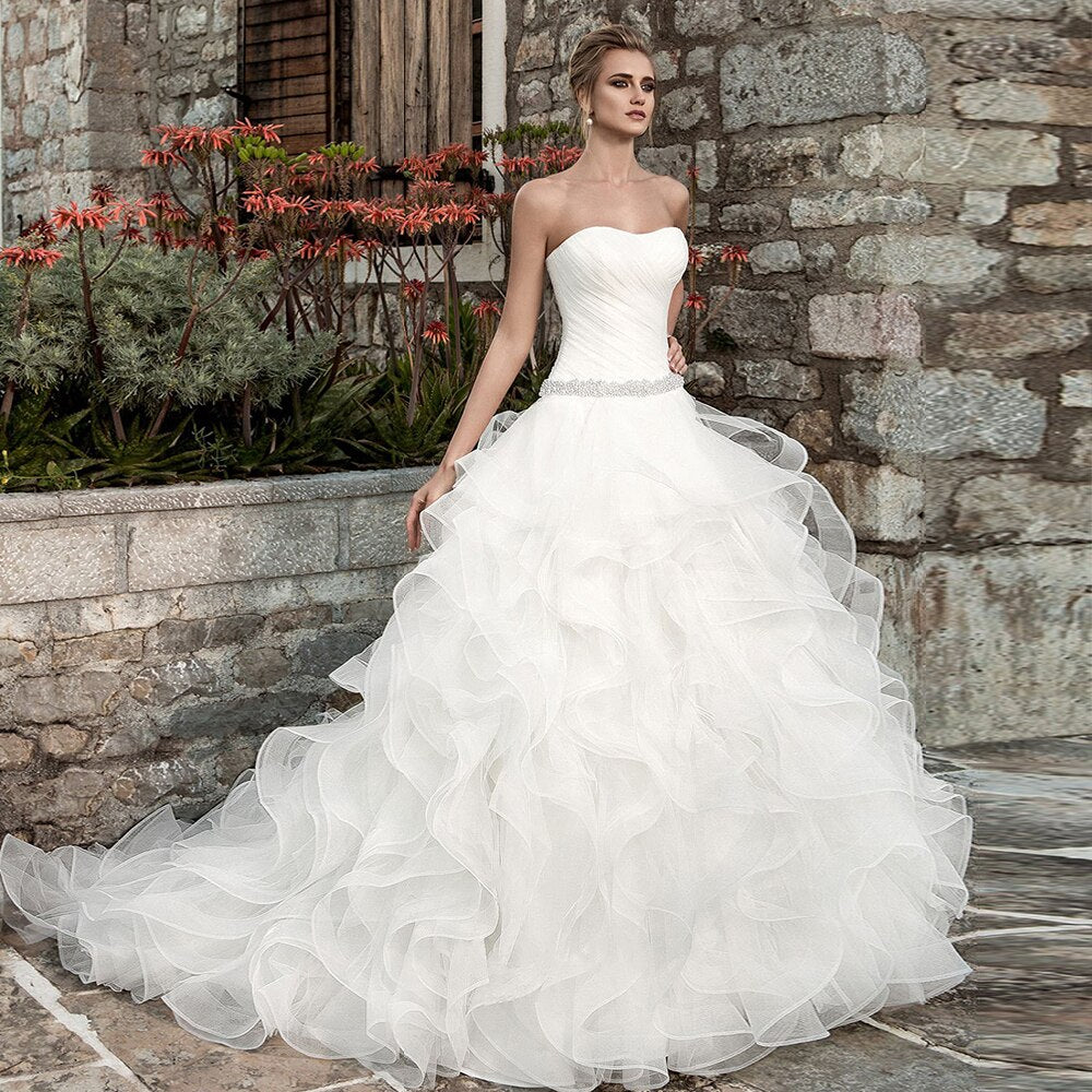 Elegant strapless princess ball gown wedding dress featuring a pleated bodice, crystal-beaded waist, and a voluminous ruffled organza skirt, set against a rustic stone backdrop.