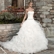 Elegant strapless princess ball gown wedding dress featuring a pleated bodice, crystal-beaded waist, and a voluminous ruffled organza skirt, set against a rustic stone backdrop.