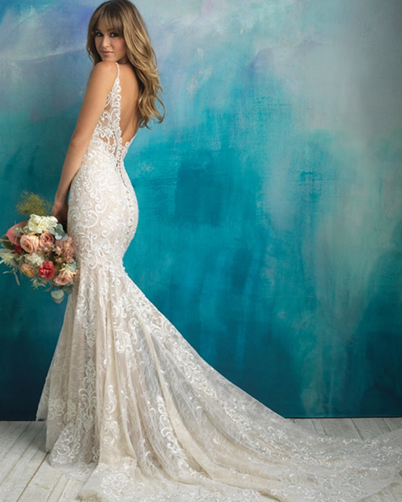 Side view of a bride in a mermaid-style wedding gown with a low backless design, lace appliques, and a long train, holding a bouquet.